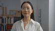 © Krakenimages.com - Chinese woman wearing glasses in an office setting, looking confident and professional inside a modern workplace, with shelves and books in the background, creating a focused environment.