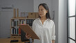 © Krakenimages.com - Woman in glasses wearing a white shirt holding a clipboard in a modern office with shelves, books, and a computer in the background.