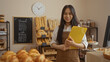 © Krakenimages.com - Young chinese woman stands in a bakery holding a clipboard, showcasing various breads and pastries with a chalkboard menu and clock in the background.