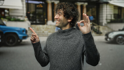 Young hispanic man pointing sideways with a cheerful expression on a city street, capturing a moment of positivity and engagement in an outdoor urban environment.