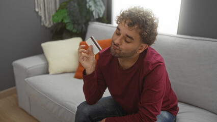 Young hispanic man sitting indoors on couch holding card with thoughtful expression in modern living room apartment setting.