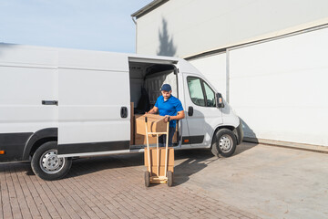 Fototapete - Delivery worker unloading boxes from van with hand truck. Courier in blue uniform unloading cardboard boxes from a delivery van using a hand truck. Logistics and shipping concept