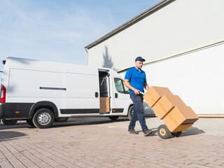 Fototapete - Delivery worker transporting boxes with a hand truck. Courier in a blue uniform moving cardboard boxes with a hand truck next to a delivery van. Logistics and shipping concept.