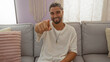 © Krakenimages.com - Young arab man sitting in a living room, smiling and pointing at the camera while wearing a white shirt, showcasing a warm and inviting home interior.