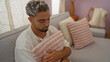 © Krakenimages.com - Young arab man sitting indoors in a cozy home holding a pink cushion, depicting comfort and relaxation in an urban apartment setting