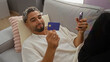 © Krakenimages.com - Young arab man holding credit card and phone while relaxing on sofa at home interior living room