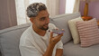 © Krakenimages.com - Young arabic man speaking into a smartphone while sitting in a cozy living room with a light-colored sofa and decor.