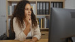 © Krakenimages.com - Young woman with curly hair working in an office environment, focusing intently on a computer screen, surrounded by shelves filled with books and binders.
