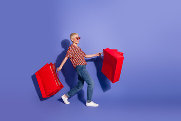 Young stylish woman in casual attire holding vibrant red shopping bags, confidently striding against a colorful purple background