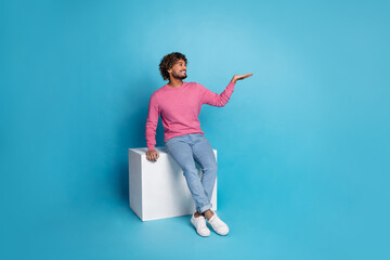 Wall Mural - Young man wearing a stylish pink jumper poses on a cube against a blue background, showcasing a friendly gesture with his hands.