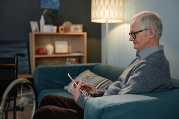 Side view of relaxed senior male wheelchair user typing message on tablet computer wearing glasses while sitting on comfortable couch in cozy living room, copy space