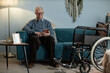 © Seventyfour - Full length shot of tech savvy grandfather using wheelchair sitting on sofa typing on digital tablet in cozy blue colored living room