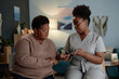 © Seventyfour - African American elderly woman with social care needs asking social worker questions about digital tablet use and online payment services, while sitting in cozy living room