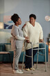 © Seventyfour - Full length shot of accommodating Black female caregiver talking to senior woman walking with frame while helping her around house providing home care for elderly, copy space
