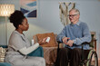 © Seventyfour - Side view of African American female social worker talking to elderly man in wheelchair explaining medication dosage holding plastic bottle of prescribed pills, while visiting patient at home