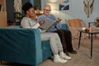 © Seventyfour - Full length of happy elderly man sitting on couch with female caregiver looking through vintage photograph album in living room during home care visit