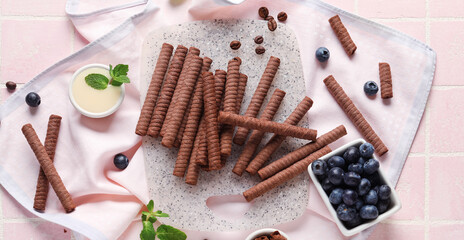 Wall Mural - Board with delicious chocolate wafer rolls, blueberries, condensed milk and coffee beans on pink tile background