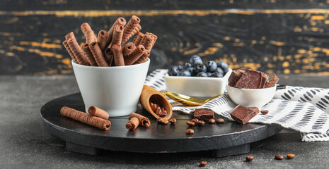 Wall Mural - Bowl with delicious wafer rolls, blueberries, chocolate pieces and coffee beans on black wooden board against wooden background