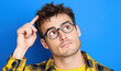 © hocine - Young man in plaid shirt and glasses looking upwards, pondering an idea or solution against a vibrant blue background, showcasing curiosity and thoughtfulness.
