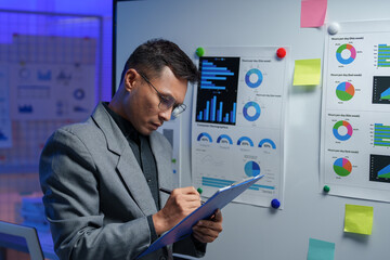 Wall Mural - Business analyst in an office setting, reviewing data charts on a whiteboard. The scene is illuminated with blue lighting, emphasizing focus on analytics and strategic planning