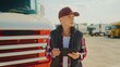 © VAKSMANV - Lorry driver standing outside in middle of parking lot. Truck behind beautiful Caucasian woman with baseball hat. Female spending free time by reading news online. Using tablet device.