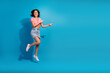 © deagreez - Energetic young woman jumping and pointing outward on a vibrant blue background