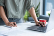 © MIND AND I - Businesswoman sitting at desk on couch in workplace or at home working on laptop and analyzing data on charts and graphs and writing on papers to make business plan and strategies for company