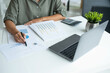 © MIND AND I - Businesswoman sitting at desk on couch in workplace or at home working on laptop and analyzing data on charts and graphs and writing on papers to make business plan and strategies for company