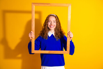 Wall Mural - Cheerful young woman holding a wooden frame against bright yellow background