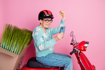 Wall Mural - Smiling Elderly Woman Wearing Helmet On Red Scooter Making Celebratory Gesture While Using Smartphone