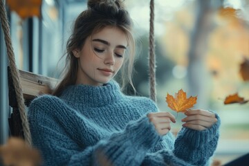 Wall Mural - A young woman in a blue sweater sits on a swing, holding a single autumn leaf.