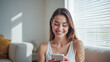© Sync - Happy young woman using smartphone at Home, smile and hold phone on her hand with internet technology use of shopping, search, chat, message at inddor living room with beautiful natural light