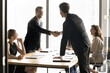 © fizkes - Two millennial businessmen shake hands standing at conference table, establish strategic alliance between two companies to collaborate on project, product development, market expansion. Contract, deal