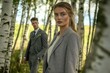 © Anjali - A woman in a grey blazer stands in a birch forest, a man in a suit blurred in the background.