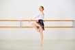 © TRAVELARIUM - Young female ballerina with brown hair in white top, black skirt, and pink tights, practices moves in bright studio. Wooden barres line neutral-toned wall