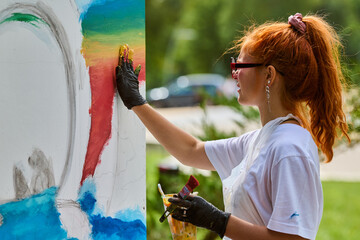 Young woman with red hair paints large canvas outdoors. Bright colors dominate, with focus on creativity and expression under natural light