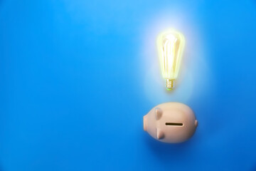 Wall Mural - Piggy bank and glowing light bulb on blue background, top view. Space for text. Energy saving concept