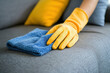 © tl6781 - hand wearing yellow rubber gloves is cleaning a gray fabric sofa with a blue microfiber cloth. The background features decorative pillows, creating a cozy and hygienic home setting