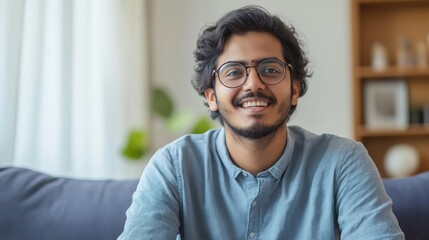 Poster - A cheerful young Indian man sits on a soft sofa, exuding warmth and positivity. Sunlight gently streams through the window, enhancing the inviting atmosphere of the living room