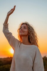 Wall Mural - A woman with long hair is standing in a field and waving her hand. The sun is setting in the background, creating a warm and peaceful atmosphere