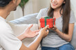 © KMPZZZ - Celebrating Valentine's day anniversary, relationship two people romantic asian young couple love, boyfriend gives a box gift to her girlfriend, happy woman getting present sitting on sofa at home.