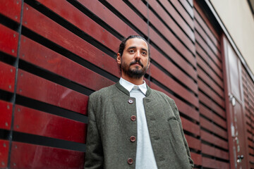 Handsome fashionable guy in stylish clothes with coat standing near red wall on street. Cool trendy man