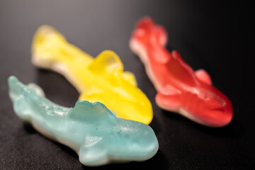 Wall Mural - A close-up of three gummy shark candies in blue, yellow and red arranged on a black background. The candies have a shiny, translucent texture and a soft, chewy appearance.