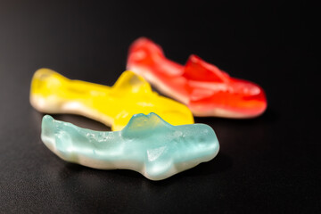 Wall Mural - A close-up of three gummy shark candies in blue, yellow and red arranged on a black background. The candies have a shiny, translucent texture and a soft, chewy appearance.