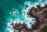 Aerial photograph of a rocky coastline with turquoise waves crashing against dark, jagged rocks, highlighting the dynamic movement and texture.

