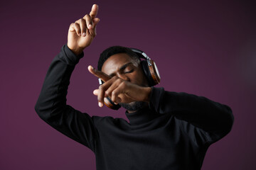 Young Black man enjoying music with headphones, dancing and expressing joy, against a vibrant purple background, showcasing a stylish and modern lifestyle