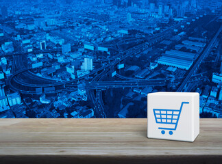 Wall Mural - Shop cart icon on white block cube on wooden table over modern city tower, street, expressway and skyscraper, Business shopping online concept