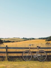 Poster - Bicycle resting against a wooden fence in a golden field under a clear sky during sunny daytime. Generative AI