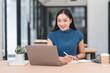 © PRIME STOCK LAB - Asian woman in blue top smiles while working on laptop in modern office. She takes notes and enjoys her work.