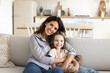 © fizkes - Here lives happiness. Portrait happy family beautiful young mommy cute little daughter cuddle on sofa in cozy kitchen interior looking at camera with candid smiles enjoy spending time at home together